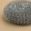 Detailopname van een roestvrijstalen staalspons vanuit een schuine hoek, waarbij de fijn gevlochten staaldraden en de stevige, duurzame structuur scherp in beeld zijn tegen een neutrale beige achtergrond.