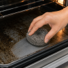 Staalspons in gebruik voor het reinigen van een ovenbakplaat met hardnekkige aangekoekte vet- en etensresten. De roestvrijstalen spons verwijdert vuil effectief en laat het oppervlak weer schoon achter.