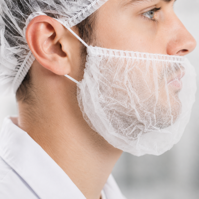 Gedetailleerde close-up van een witte disposable baardcover die achter de oren wordt bevestigd met elastische lussen. De afbeelding toont de fijne non-woven vezelstructuur, de elastische zoom en de comfortabele pasvorm rond de kaaklijn. De baardcover bede