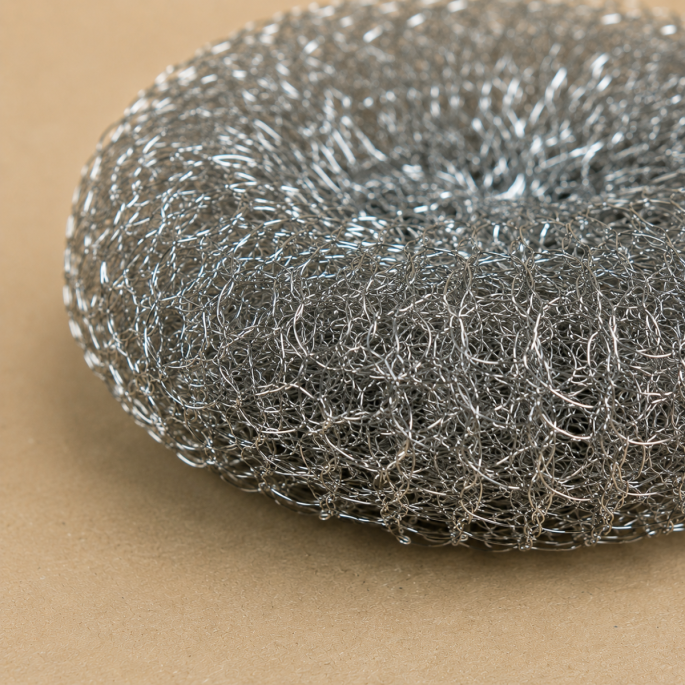 Detailopname van een roestvrijstalen staalspons vanuit een schuine hoek, waarbij de fijn gevlochten staaldraden en de stevige, duurzame structuur scherp in beeld zijn tegen een neutrale beige achtergrond.