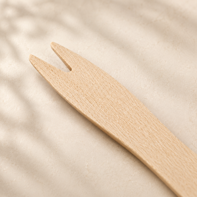 Detailfoto van een houten amusevorkje van licht berkenhout, schuin gefotografeerd op een zachte beige achtergrond. De close-up toont de fijne houtnerf, de tweepuntige vorkvorm en de natuurlijke afwerking van het duurzame wegwerpbestek.