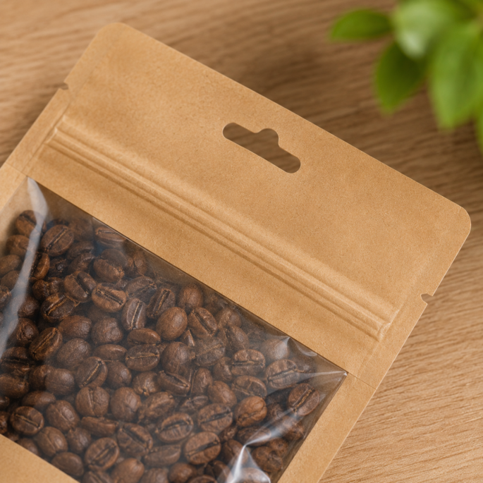Detailopname van een kraft papieren stazak met transparant venster, gevuld met koffiebonen. De close-up toont het kijkvenster, de hoogwaardige afwerking en het ophanggat van de verpakking, waardoor de inhoud duidelijk zichtbaar is.