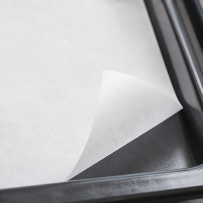 Close-up detailfoto van wit gesiliconeerd bakpapier in een zwarte bakplaat, met omgevouwen hoek die de fijne textuur, gladde anti-aanbaklaag en premium kwaliteit van het papier benadrukt.