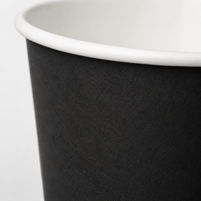 Extreme close-up van een zwarte disposable papieren beker, waarbij de fijne materiaalstructuur, matte afwerking en witte bovenrand van de beker duidelijk zichtbaar zijn.